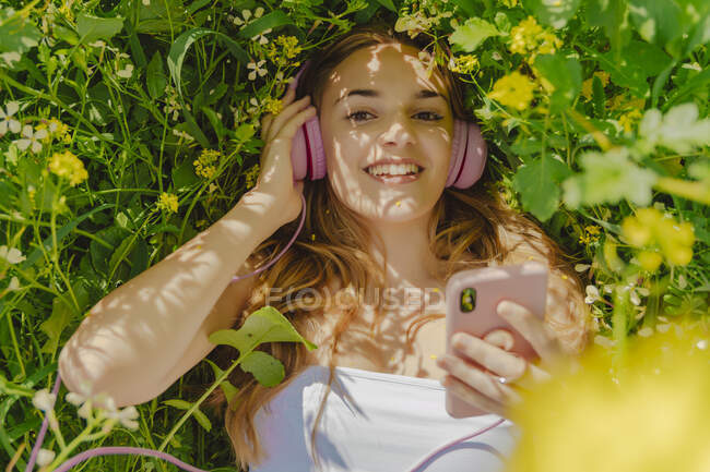 Retrato de una joven feliz con auriculares y teléfono inteligente que yace en un prado de flores en primavera - foto de stock