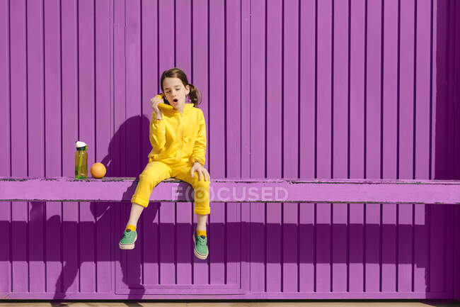 Маленькая девочка в желтом сидит на баре перед фиолетовым фоном играя с бананом — стоковое фото