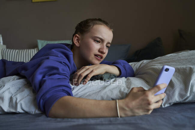 Retrato de menina deitada na cama em casa usando smartphone para vídeo chat — Fotografia de Stock