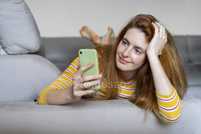 Mujer joven acostada en el sofá de su casa con un smartphone - foto de stock