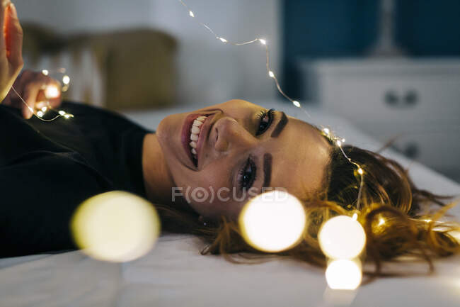 Ritratto di giovane donna felice sdraiata a letto con catena di luci — Foto stock