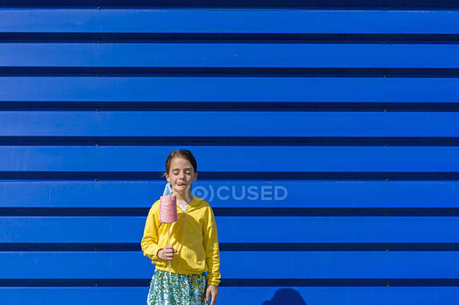 Little girl with fake ice cream standing in front of blue background licking lips — Stock Photo