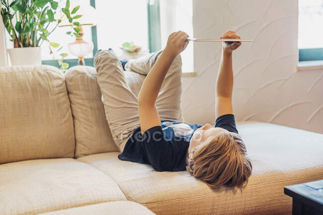Boy lying on couch in living room at home using tablet — Stock Photo