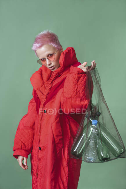 Портрет молодой женщины с короткими розовыми волосами в красном пальто, держащей сумку с пластиковыми бутылками — стоковое фото