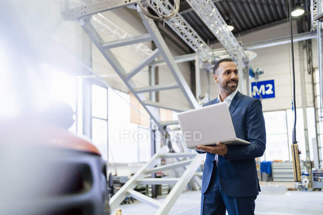 Homme d'affaires souriant tenant un ordinateur portable dans une usine — Photo de stock