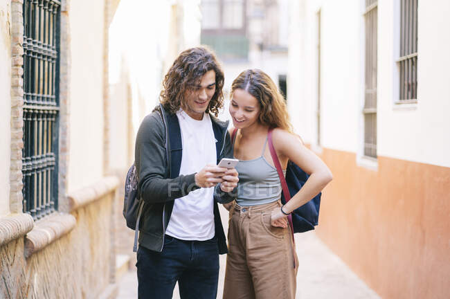 Mujer joven sonriente mirando al hombre usando un teléfono inteligente mientras está de pie en la calle estrecha en Santa Cruz, Sevilla, España - foto de stock