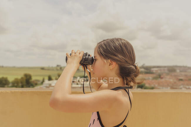 Chica en traje de baño de pie en la terraza de la azotea mirando a través de binoculares - foto de stock