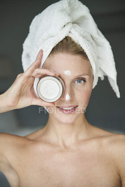 Porträt einer schönen Frau mit einem in ein Handtuch gehüllten Kopf, der ein Cremeglas vor ihrem Auge hält — Stockfoto