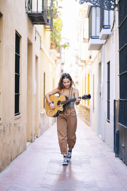 Intera lunghezza della giovane donna che suona la chitarra mentre cammina in una strada stretta tra edifici a Santa Cruz, Siviglia, Spagna — Foto stock