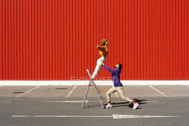 Joven hombre y mujer actuando con una escalera delante de una pared roja - foto de stock