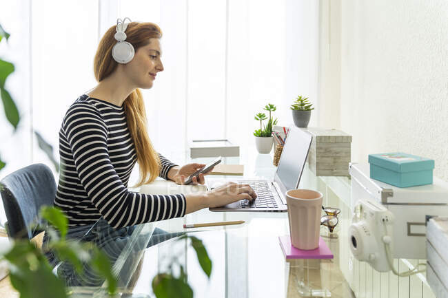 Young woman wearing headphones using laptop and smartphone at desk at home — Stock Photo
