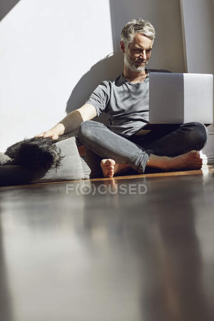 Зрілий чоловік сидить вдома на підлозі за допомогою ноутбука і гладить собаку. — стокове фото