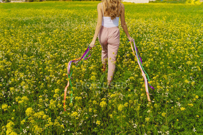 Visão traseira de mulher jovem que anda com fitas coloridas em um prado de flor na primavera — Fotografia de Stock