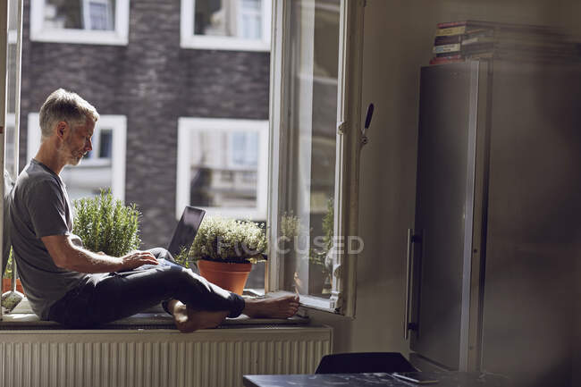 Hombre maduro sentado en la ventana en casa usando el ordenador portátil - foto de stock