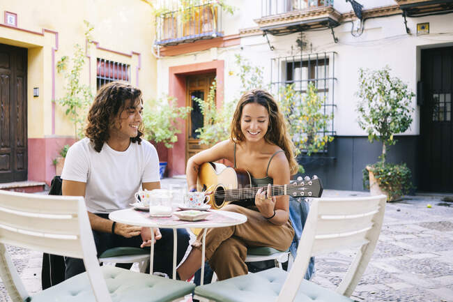Усміхаючись, молодий чоловік дивиться на дівчину, яка грає на гітарі, сидячи в кафе на тротуарі в Санта - Крус (Севілья, Іспанія). — стокове фото