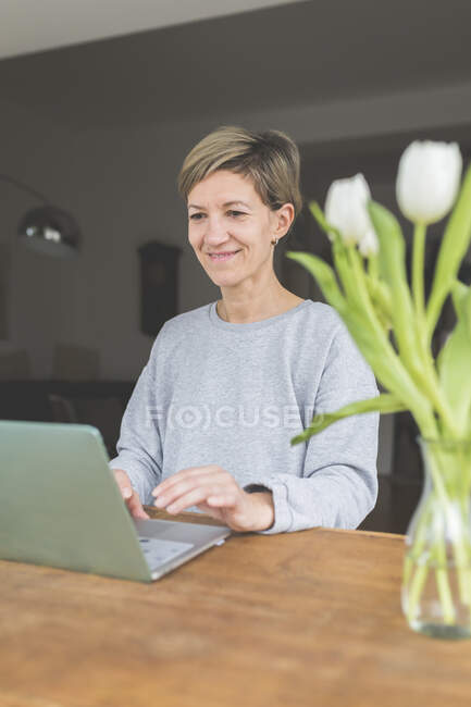 Mature woman working from home, using laptop on table with flowers — Stock Photo