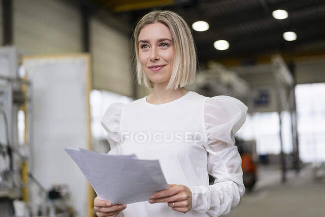 Porträt einer lächelnden jungen Frau mit Papieren in einer Fabrik — Stockfoto