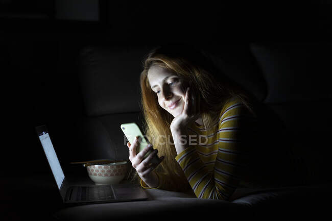 Молодая женщина лежит на диване дома с помощью ноутбука и смартфона — стоковое фото