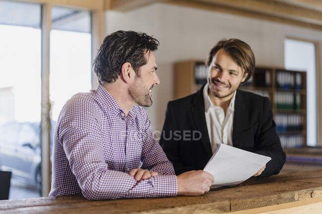 Два улыбающихся бизнесмена с документами разговаривают в деревянном офисе открытой планировки — стоковое фото