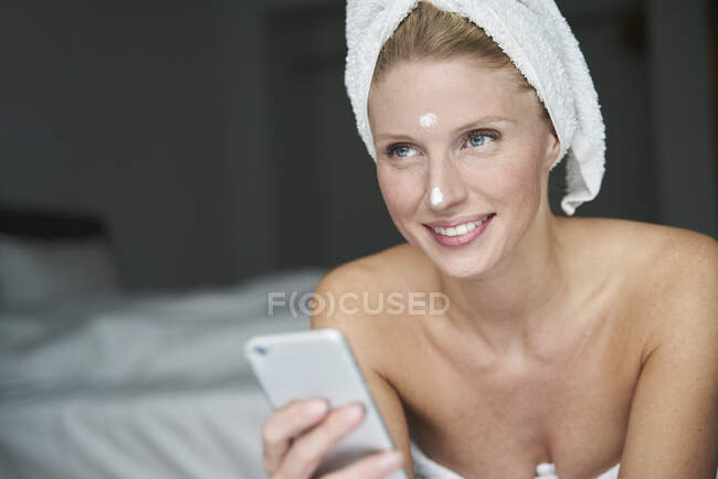 Retrato de una hermosa mujer con la cabeza envuelta en una toalla que sostiene el teléfono inteligente - foto de stock