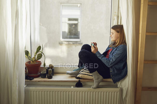 Ragazza seduta alla finestra a casa con smartphone — Foto stock