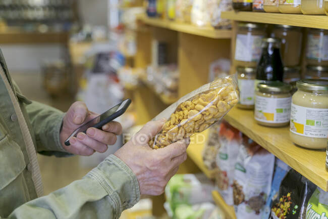 Зблизька старший чоловік з смартфоном купує продукти в маленькому продовольчому магазині перевіряє інгредієнти — стокове фото