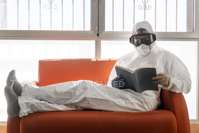 Mujer con ropa protectora, sentada en el sofá, leyendo libro - foto de stock