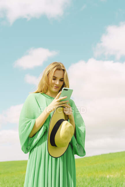 Молодая женщина в зеленом платье стоит на поле с помощью смартфона — стоковое фото