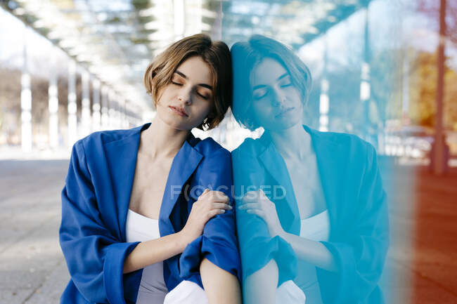 Jeune femme aux yeux fermés appuyée sur un mur de verre coloré avec son reflet — Photo de stock