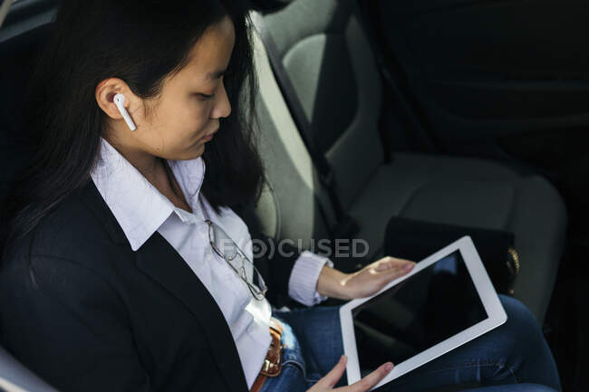 Young businesswoman sitting in car using earpods and digital tablet — Stock Photo