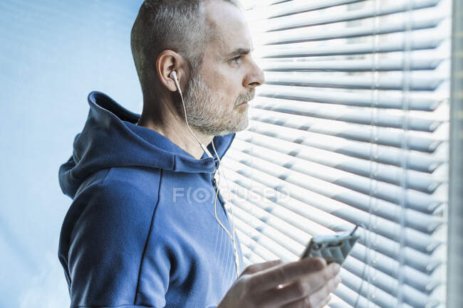 Uomo pensieroso con smartphone e auricolari guardando fuori dalla finestra veneziana cieca — Foto stock