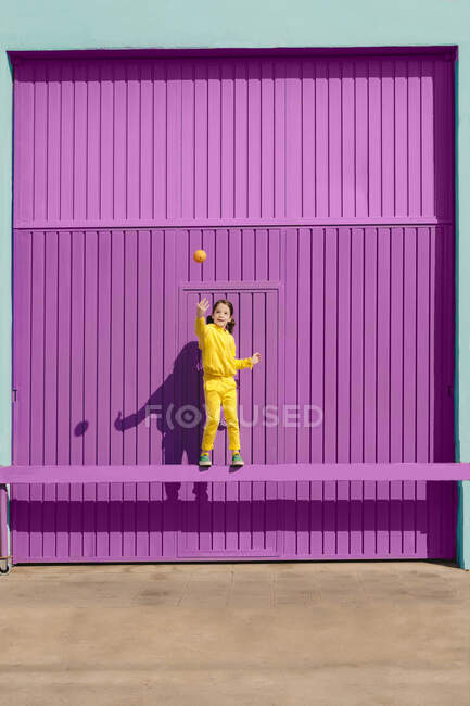 Щаслива дівчинка в жовтому одязі, що балансує на барі перед фіолетовим гаражем, що кидає апельсин в повітря. — стокове фото