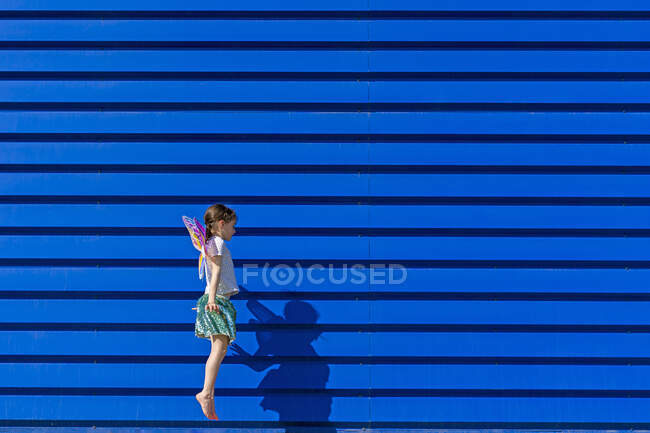 Kleines Mädchen mit bunten Schmetterlingsflügeln springt vor blauem Hintergrund in die Luft — Stockfoto