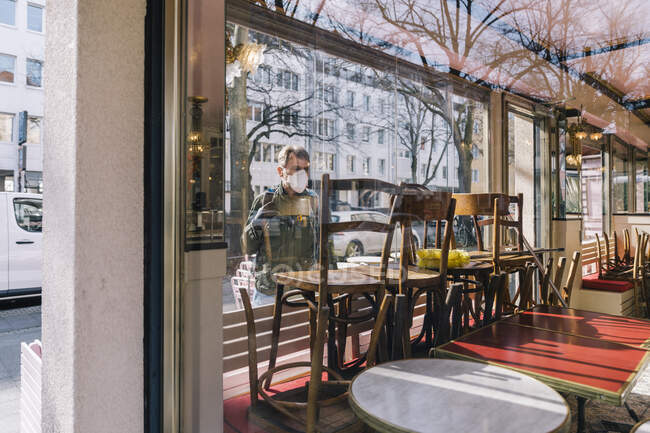 Человек в маске смотрит через окно в закрытый ресторан — стоковое фото