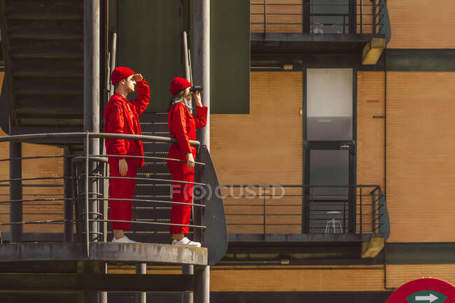 Молода пара в червоних накладках і капелюхах стоїть на платформі, дивлячись на відстань. — стокове фото