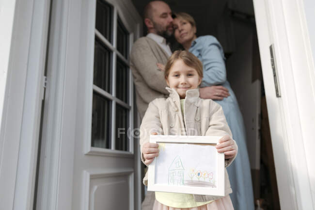 Счастливая маленькая девочка с рисунком ее дома и родители обнимаются за ней — стоковое фото