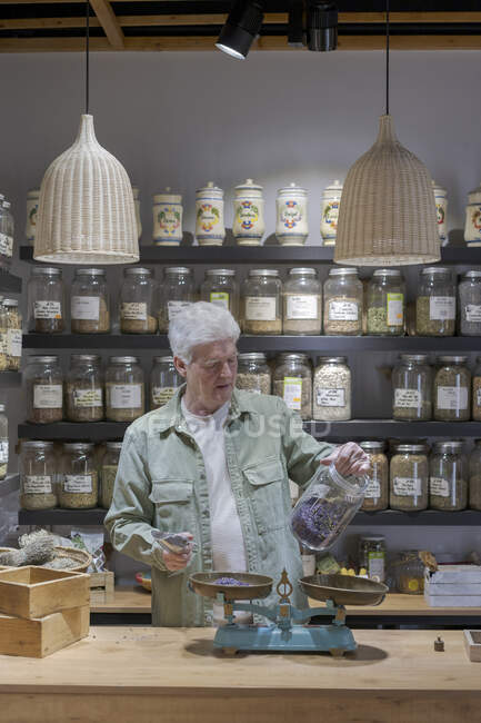 Senior man behind the counter in his shop holding jar of dried lavender blossoms — Stock Photo