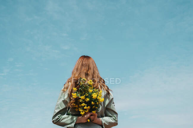 Vue arrière de jeune femme rousse contre le ciel tenant un bouquet de fleurs derrière son dos — Photo de stock