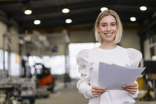 Портрет улыбающейся молодой женщины с бумагами на фабрике — стоковое фото