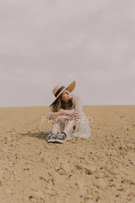 Жінка з солом'яним капелюхом і вінтажне плаття, що сидить на віддаленому полі в сільській місцевості — стокове фото