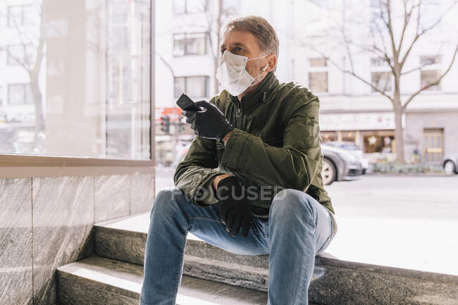Homem com máscara sentado em escadas na cidade segurando smartphone — Fotografia de Stock