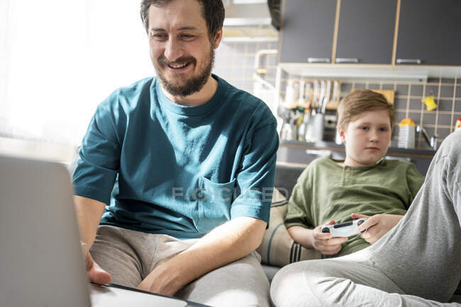 Portrait de père souriant utilisant un ordinateur portable à la maison tandis que son fils ennuyeux en arrière-plan — Photo de stock