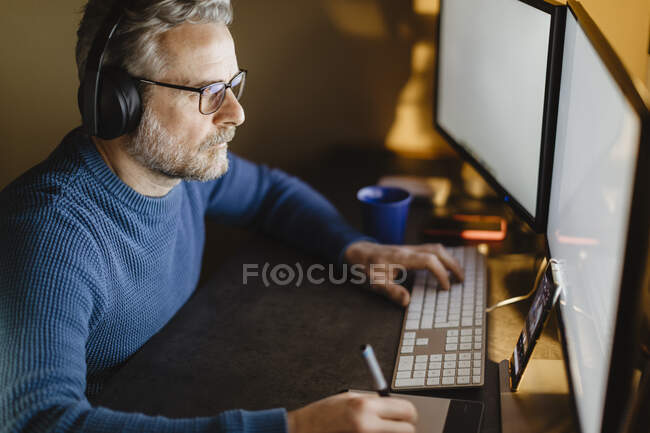 Reifer Mann mit Kopfhörern sitzt zu Hause am Schreibtisch und arbeitet mit Grafik-Tablet und Computer — Stockfoto