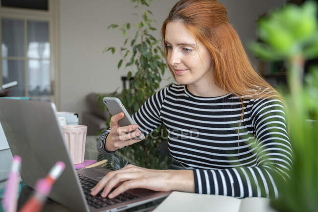 Mujer joven usando el ordenador portátil y el teléfono inteligente en el escritorio en casa - foto de stock