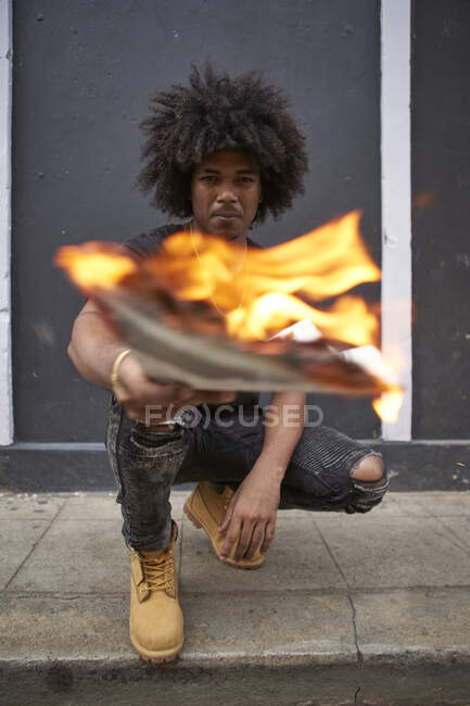 Ritratto di giovane uomo accovacciato sul marciapiede con giornale in fiamme — Foto stock