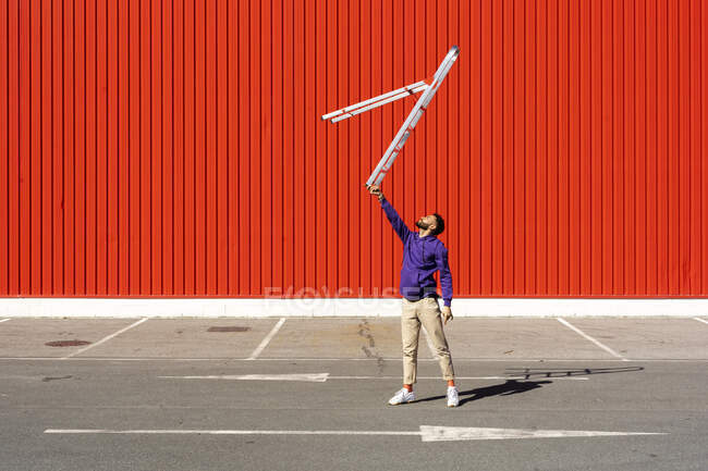 Jeune homme équilibrant une échelle devant un mur rouge — Photo de stock