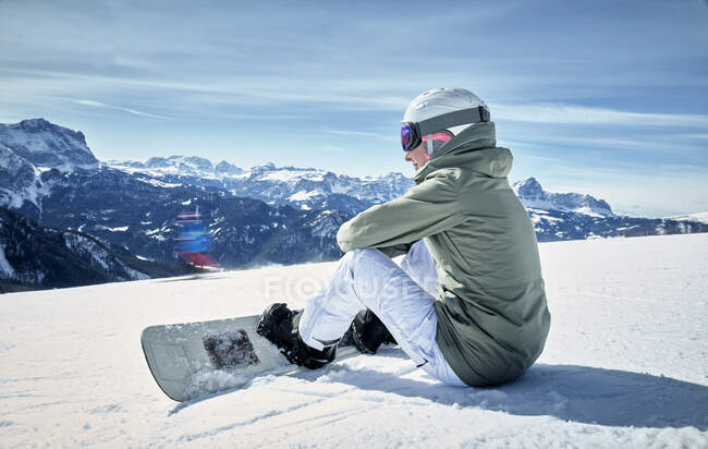 Улыбающаяся женщина сидит с сноубордом на смотровой площадке и наслаждается видом на расстоянии — стоковое фото