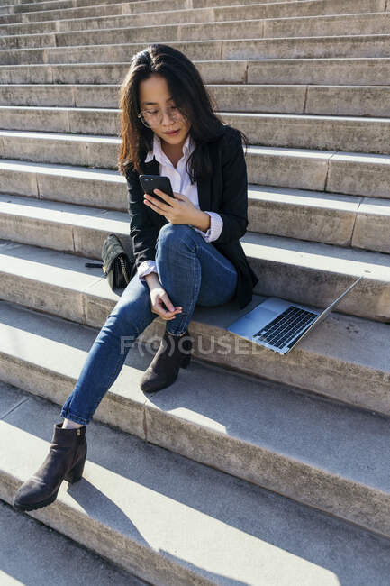 Young businesswoman with laptop sitting on stairs outdoors looking at mobile phone — Stock Photo