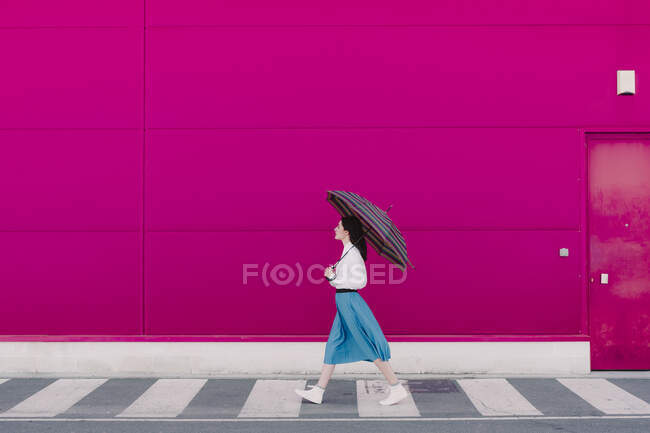 Молода жінка з парасолькою, що йде перед рожевою стіною. — стокове фото