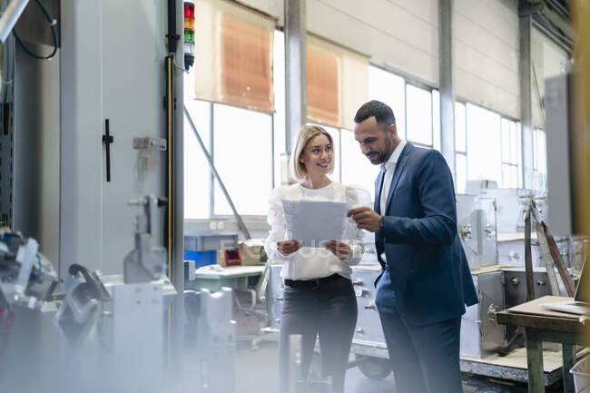 Businessman and young woman with papers talking at a machine in a factory — Stock Photo